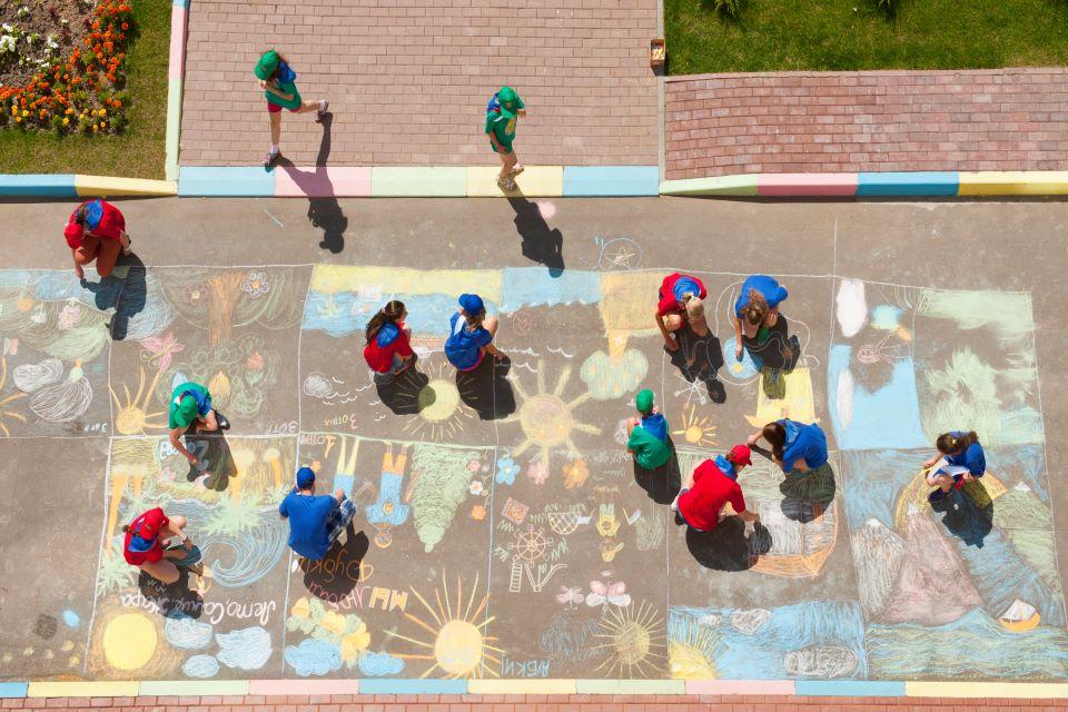 Vue du ciel sur une cour de récréation. Des enfants dessinent sur le sol à la craie.