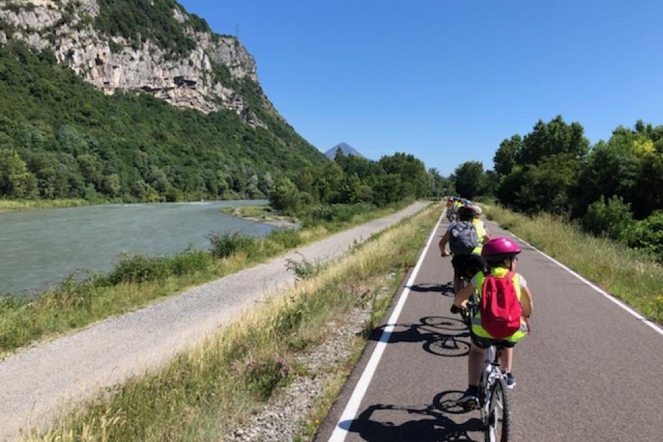 Plusieurs enfants roulent à vélo sur une piste cyclable.  La photo est prise depuis la fin de la file. Tous les enfants portent des gilets jaunes par mesure de sécurité.