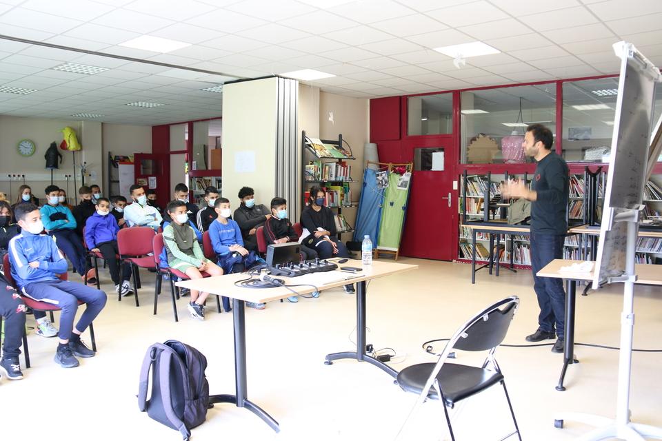 Marien Tillet fait face à une classe d'élèves. Les jeunes sont assis sur des chaises, Marien se tient debout face à eux. 