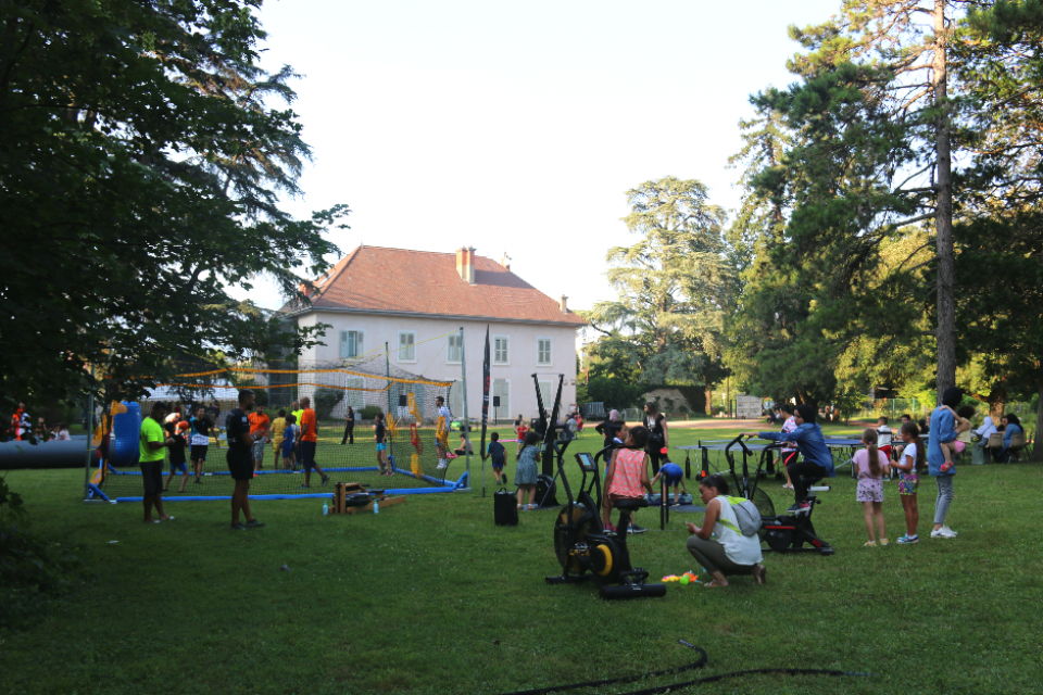 Vue d'ensemble du parc Géo-Charles pour destination été 2021. Des appareils de sports, des terrains de sports en mousse (boxe, hand) sont installés dans l'herbe. Des tables et des chaises sont installées pour les ateliers artistiques (écriture, peinture) etc. Les habitant-es sont nombreux-ses à participer.